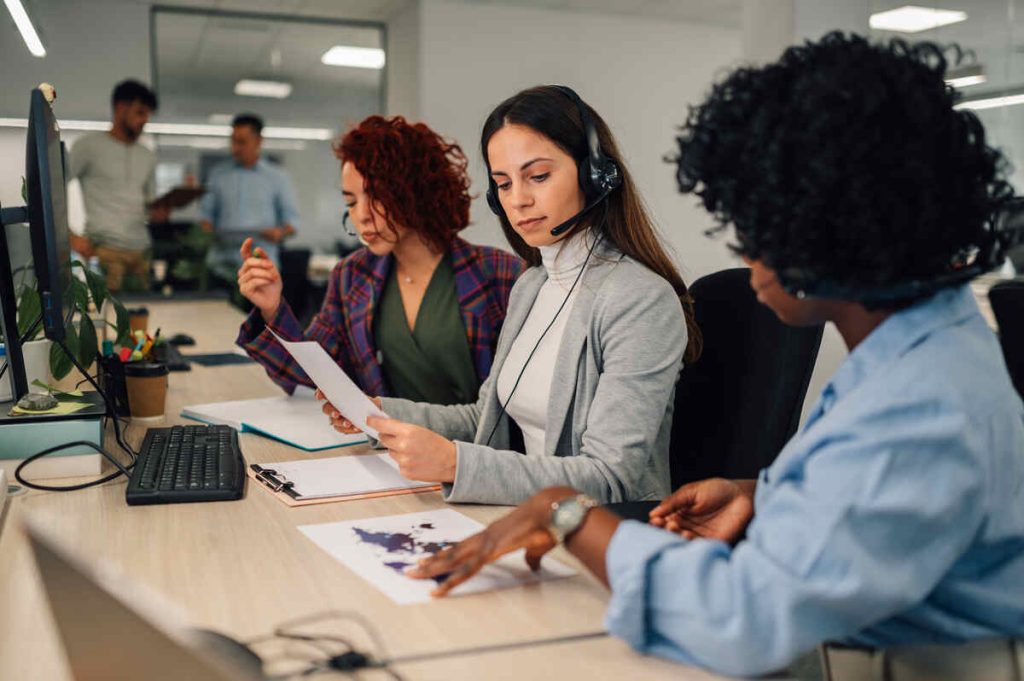 Caucasian woman with headset working at the desk and talking with clients. Call center operator with wireless headset talking with her african american colleague showing her charts. Copy space.