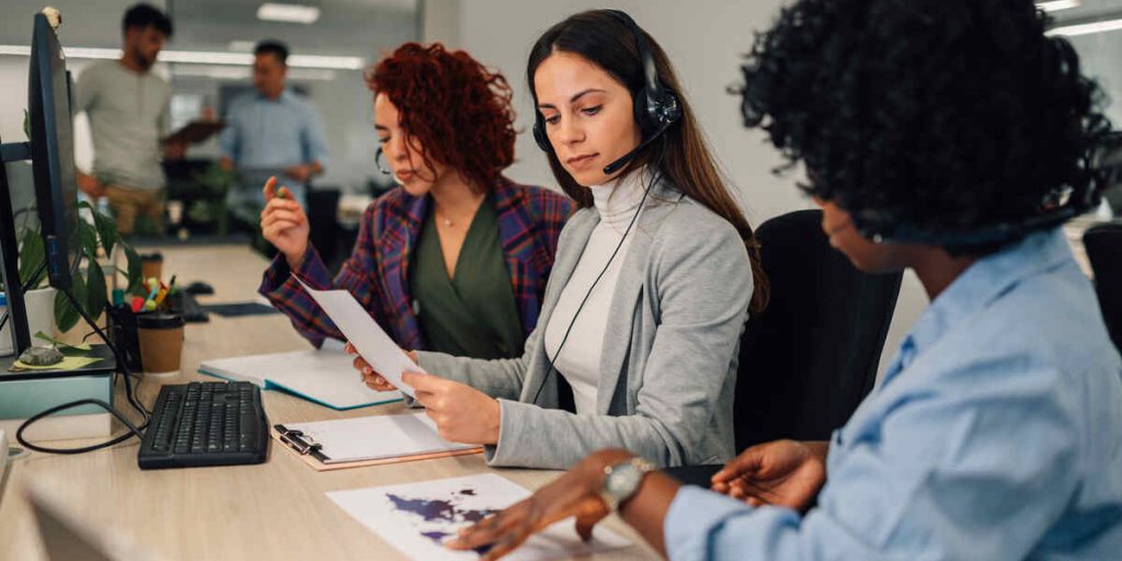 Caucasian woman with headset working at the desk and talking with clients. Call center operator with wireless headset talking with her african american colleague showing her charts. Copy space.