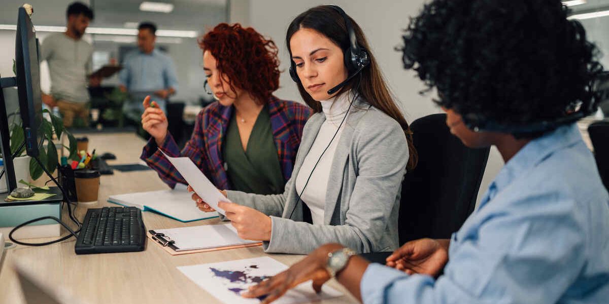 Caucasian woman with headset working at the desk and talking with clients. Call center operator with wireless headset talking with her african american colleague showing her charts. Copy space.