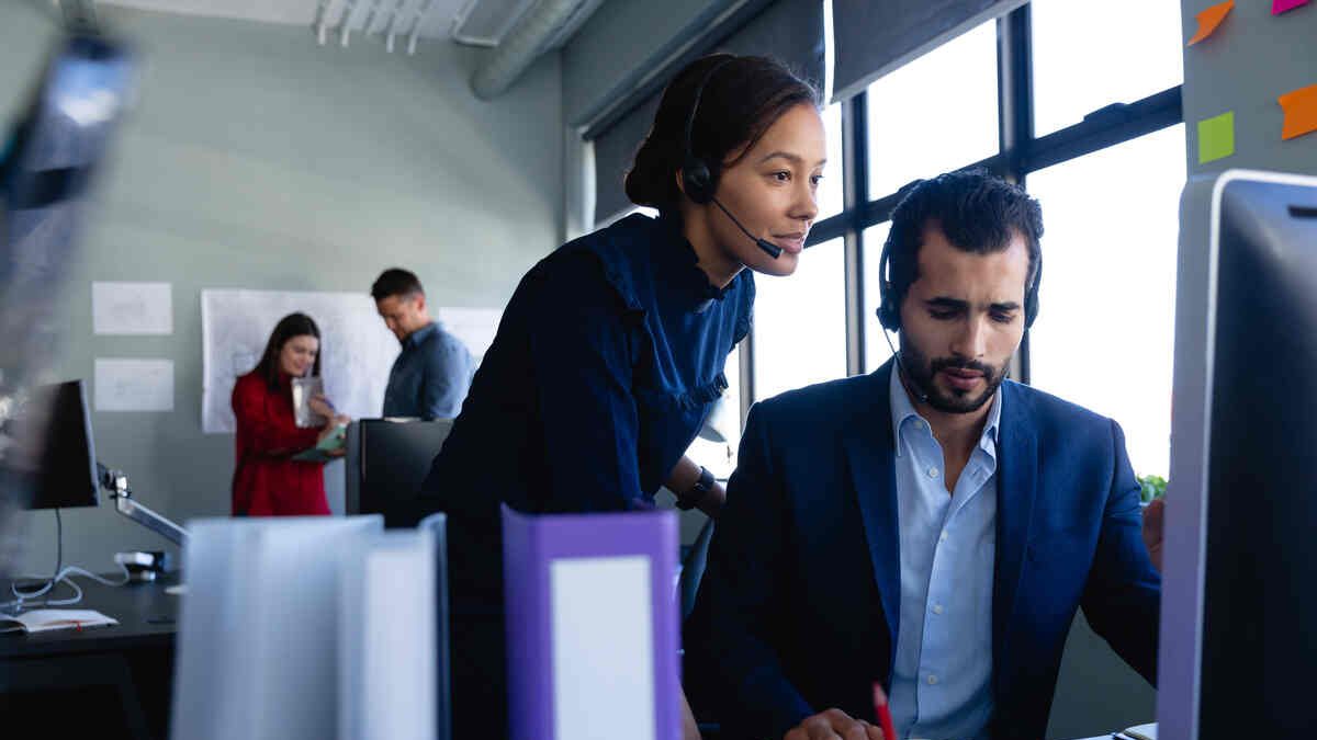 Caucasian man and mixed race woman wearing headset, using a desktop computer, with other colleagues working in the background.